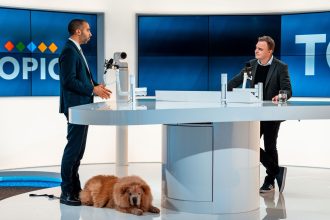 Duas pessoas conversam em um estúdio moderno de televisão, enquanto um cachorro permanece deitado ao lado.