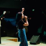 Cantora se apresenta ao vivo diante do público, com um braço levantado e um microfone à frente.